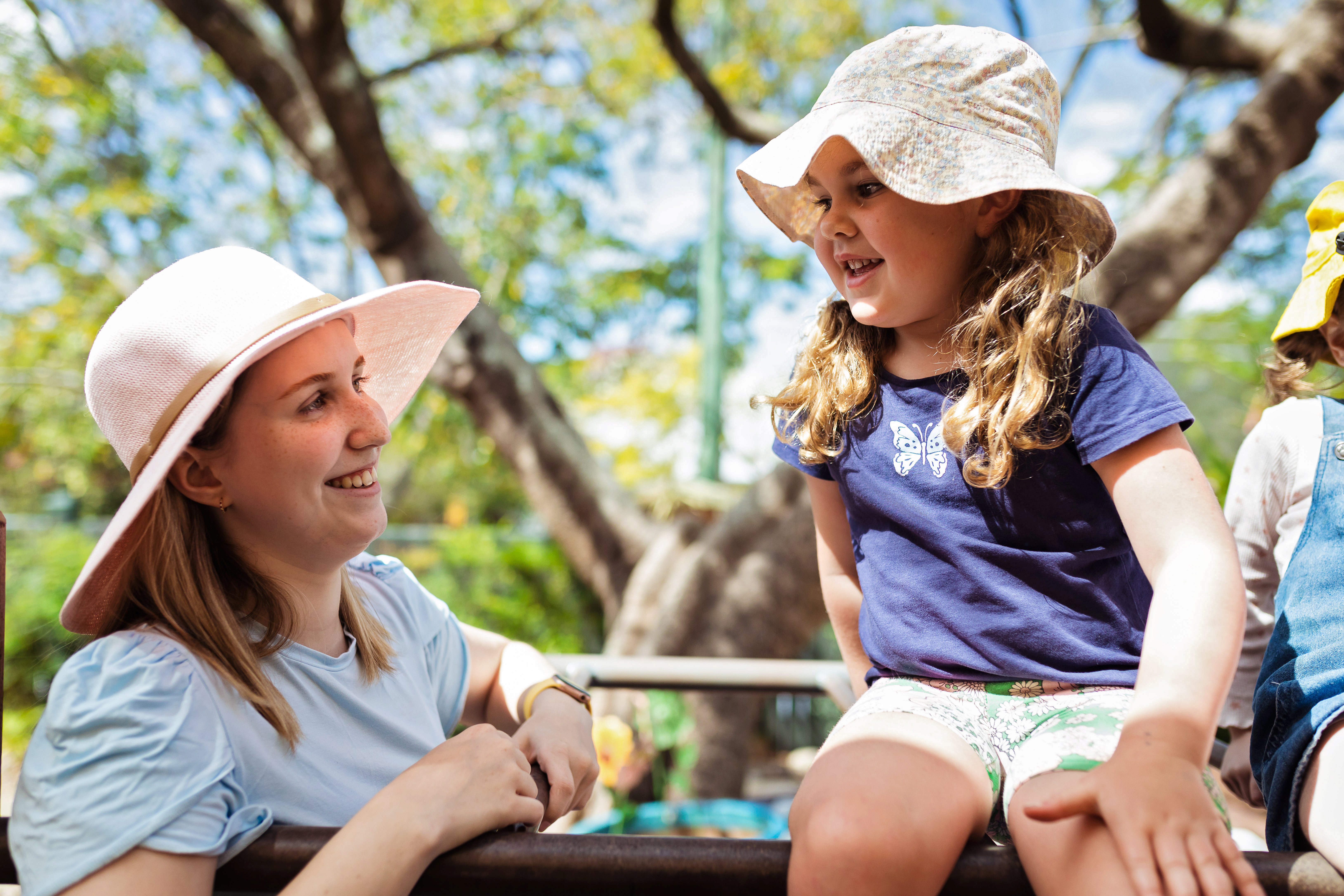 Child wearing hat outside sitting on monkey bars looking down at parent standing wearing a hat