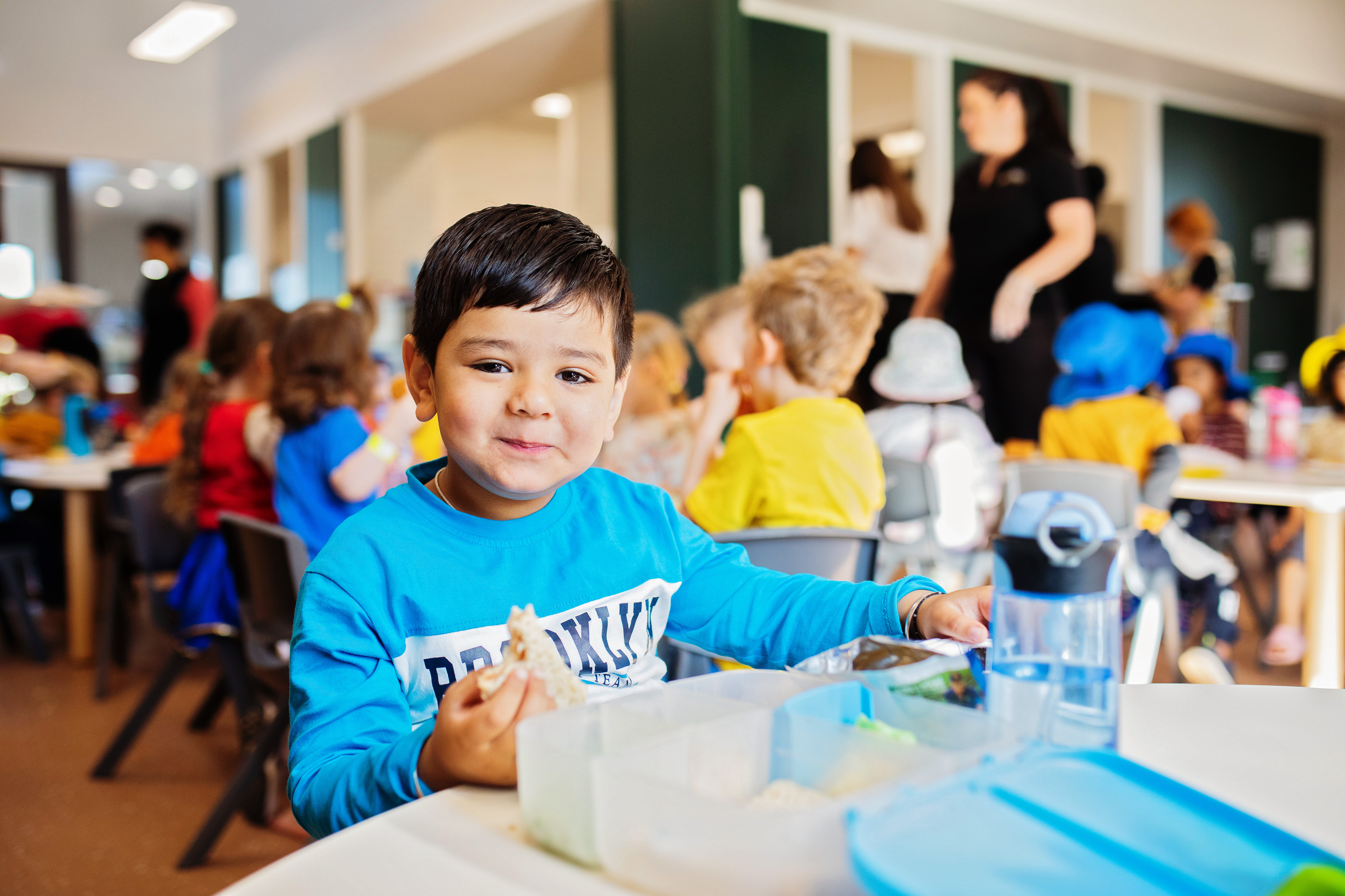 Child sitting at low table looking at camera smiling while eating morning tea