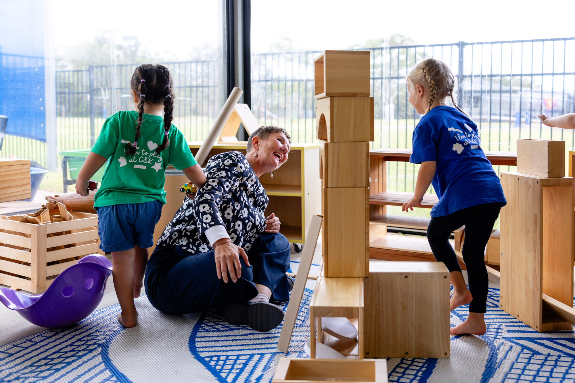 C&K Banksia Beach - children and an educator happily stacking large wooden blocks on an enclosed verandah