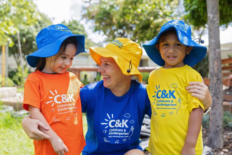 Educator outside with two kindy children in bright C&K tshirts and hats