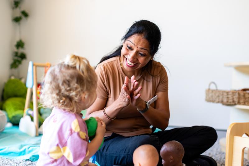 Educator smiling, clapping and singing with toddler on the floor