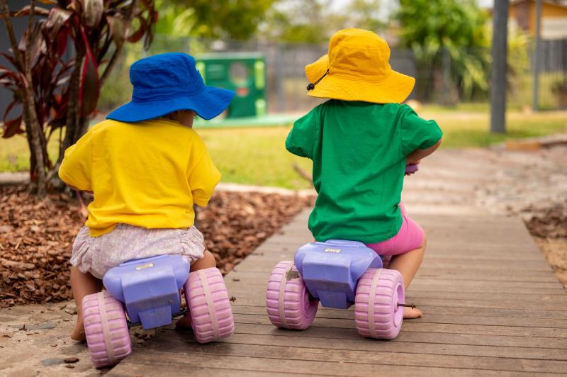 Two toddlers riding off together on trikes