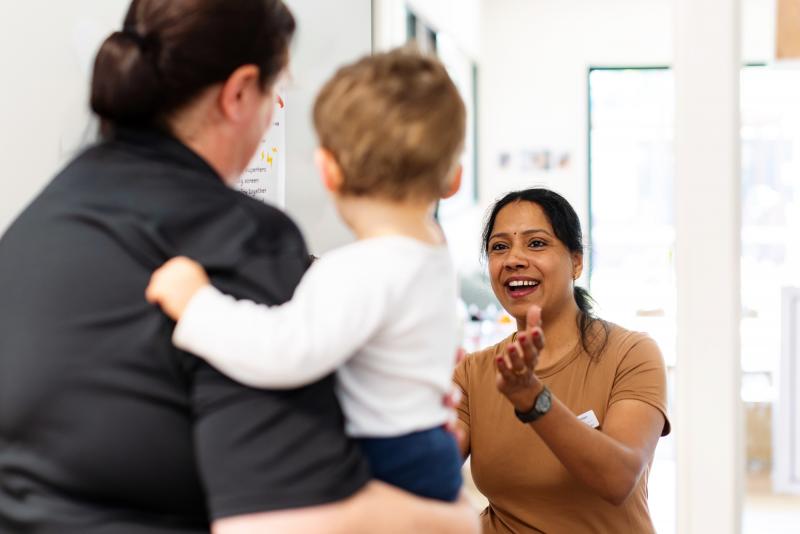 Educator smiling as a parent passes their toddler over to her