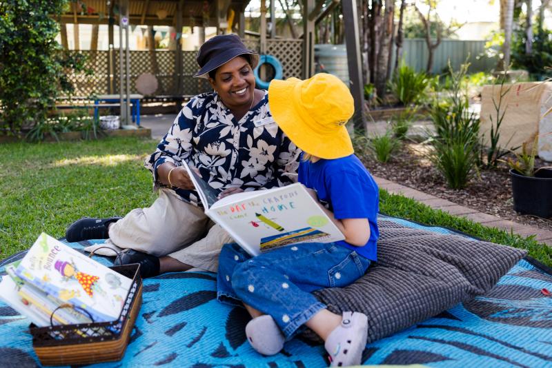 Educator reading and smiling with kindy child outside